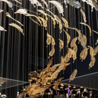 A large custom lighting hotel chandelier featuring suspended golden feather-like elements. This big chandelier showcases intricate glass pieces resembling delicate feathers in shimmering gold and clear tones. The feathers cascade downward, creating a mesmerizing waterfall effect against a dark background. This unique custom lighting design transforms the space into an elegant, ethereal environment, perfect for luxury hotel lobbies or grand ballrooms.