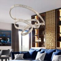 A large chandelier featuring custom lighting design with intertwining crystal-like ribbons. This big hotel chandelier showcases a fluid, sculptural form composed of transparent elements that catch and reflect light. The custom lighting piece creates graceful curves and loops, suspended above a luxurious living area with blue velvet sofas. It contrasts beautifully with the warm wood tones and geometric patterns of the surrounding decor. This spectacular chandelier serves as a captivating focal point, blending modern artistry with functional lighting in an upscale hotel space.