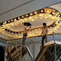 A large custom lighting installation featuring an oval-shaped chandelier integrated into a ceiling design. This hotel chandelier showcases numerous illuminated, organic-shaped glass elements arranged along the oval's perimeter, emitting a warm golden glow. The big chandelier is complemented by a series of small spotlights lining the inner edge. Two workers on scaffolding are visible, suggesting the chandelier's impressive scale. This unique lighting fixture transforms the ceiling into a stunning focal point, perfect for enhancing the ambiance of luxurious spaces like hotel lobbies or grand halls.