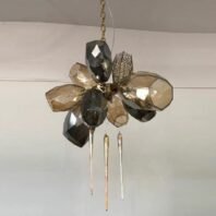 A unique and artistic pendant light showcasing innovative custom lighting design. The fixture resembles an abstract flower, with large geometric "petals" made of faceted glass or crystal in smoky gray and amber tones. These angular elements are arranged in a circular pattern around a central metallic hub, creating a sculptural, floral-inspired form. A delicate gold-toned chain suspends the pendant from the ceiling. Adding to the design's intrigue are three slender, elongated elements hanging beneath the main structure, reminiscent of pistils or stamens. One petal features a more intricate, filigree-like pattern, contrasting with the solid forms of the others. This striking custom lighting piece would serve as a captivating focal point in modern, high-end interiors, perfect for those seeking a blend of natural inspiration and contemporary geometric aesthetics.