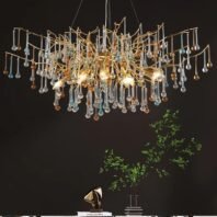 A luxurious chandelier featuring an intricate gold-toned branch structure adorned with numerous crystal droplets in various colors. The chandelier has an organic, tree-like design with golden branches extending outward and downward. Hanging from these branches are crystal pendants in clear, light blue, and amber hues, creating a multi-colored sparkling effect. The fixture is illuminated from within, with visible warm light bulbs nestled among the branches. Below the chandelier, a dark surface is partially visible, showcasing a small decorative golden object and a vase with green foliage, adding a touch of nature to the elegant setting. The chandelier is photographed against a dark background, emphasizing its radiant and intricate design.